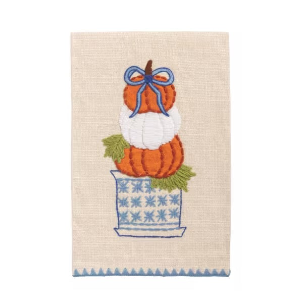 A rectangular woven kitchen towel with a stitched autumn design of stacked pumpkins in orange and white, a blue bow, green leaves, and a blue patterned pot on a cream background.