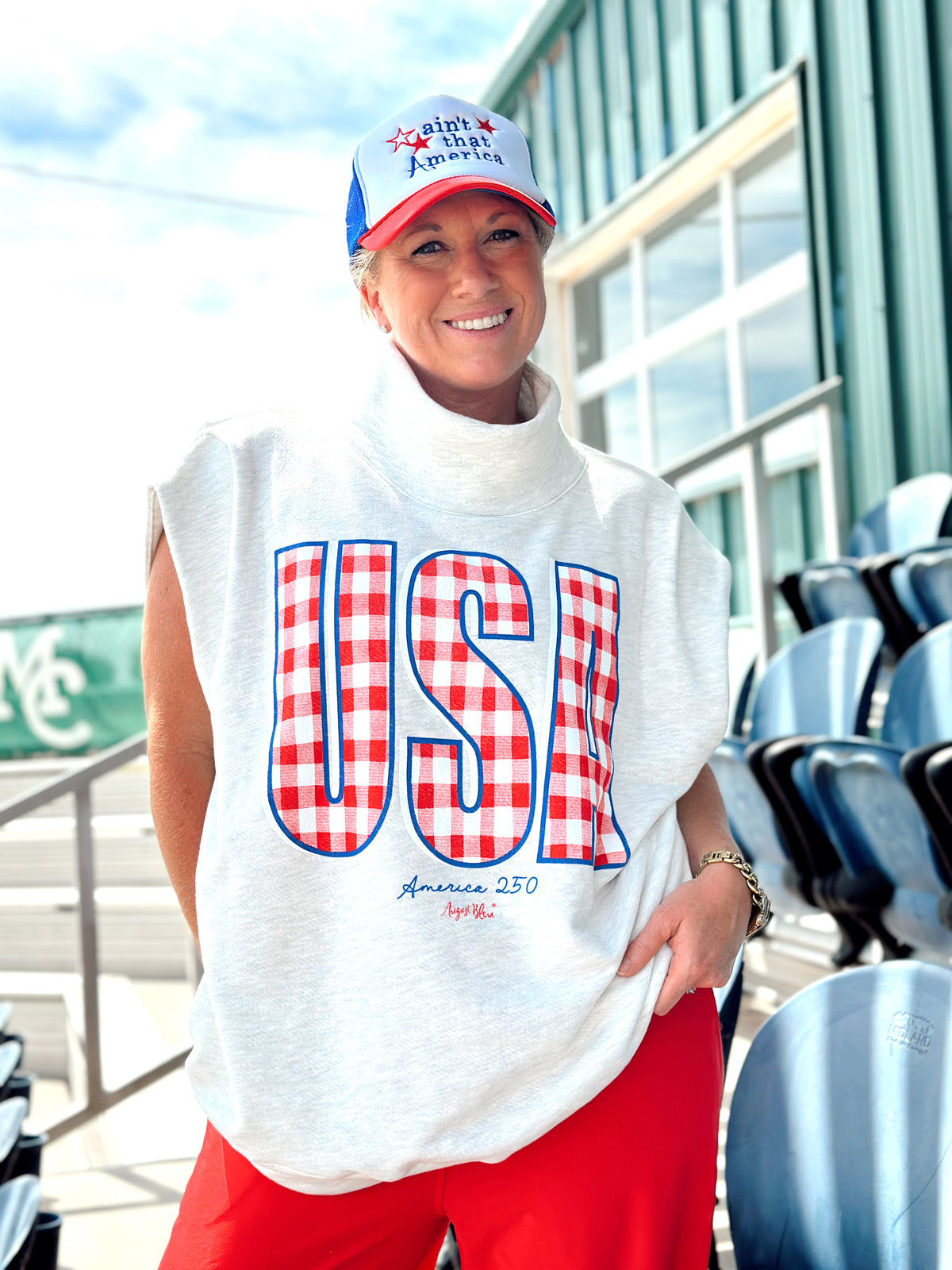 Person wearing a white sleeveless shirt with 'USA' design, red pants, and a cap outdoors.